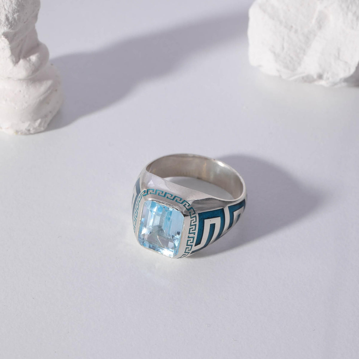 May include: A silver ring with a large, rectangular, light blue gemstone. The ring features a dark blue and white Greek key design on the sides. The ring is set against a white background.