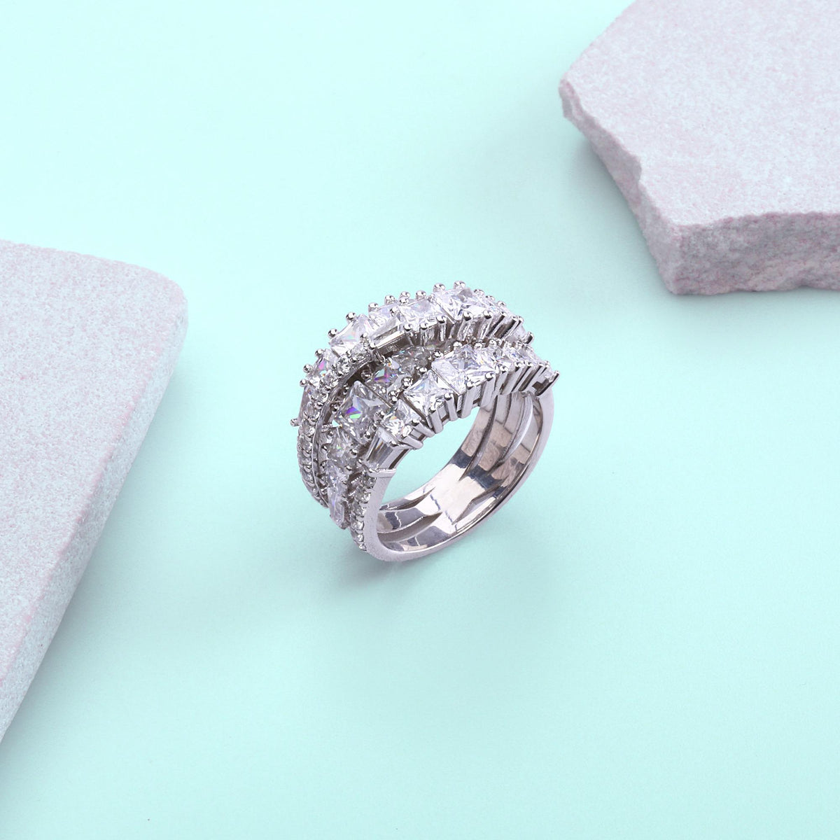 May include: A silver ring with three bands, each encrusted with square and round clear gemstones. The ring is set against a light blue background with two gray, stone-like shapes.