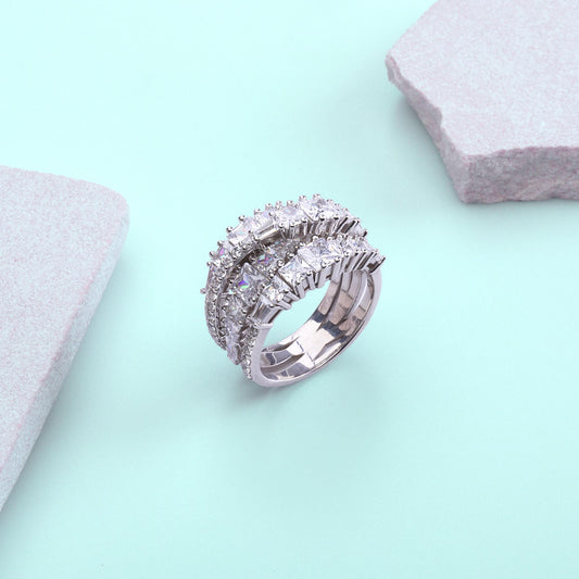 May include: A silver ring with three bands, each encrusted with square and round clear gemstones. The ring is set against a light blue background with two gray, stone-like shapes.