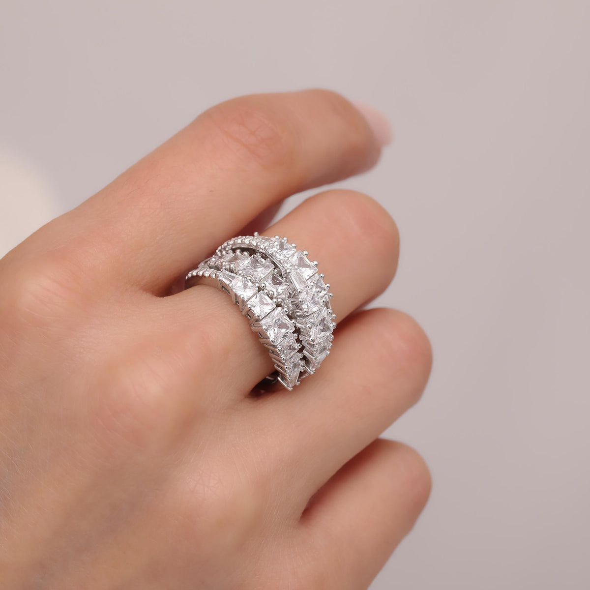 May include: A silver ring with multiple rows of square-cut, clear gemstones. The ring is worn on a finger, showcasing its intricate design and sparkling appearance. The ring's band is also detailed with smaller, round stones.